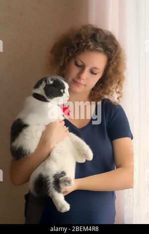 Ein Mädchen hält eine flauschige Katze in einem Schal, der aus dem Fenster schaut Stockfoto