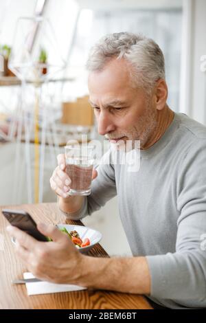 Bild eines älteren grauhaarigen bärtigen Geschäftsmann sitzen im Café essen Salat mit Handy. Stockfoto
