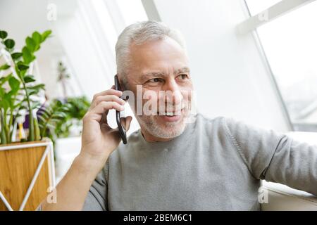 Bild eines glücklichen positiven reifen älteren grauhaarigen bärtigen Geschäftsmann sitzen im Café mit Handy. Stockfoto