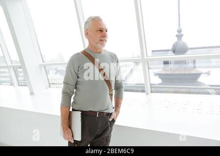 Bild eines älteren positiven grauhaarigen bärtigen Geschäftsmann in Innenräumen mit Laptop-Computer. Stockfoto