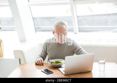 Bild eines älteren grauen-behaarten Geschäftsmannes sitzen im Café und essen mit einem Laptop. Stockfoto