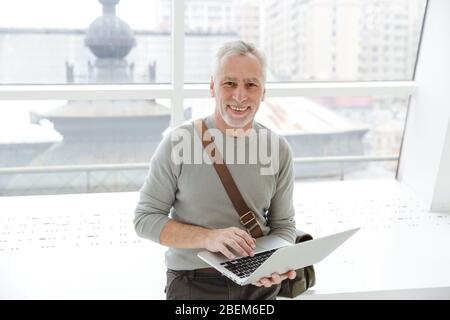 Foto eines lächelnden, grauhaarigen Mannes, der den Laptop hält und benutzt, während er drinnen in der Nähe von Fenstern steht Stockfoto
