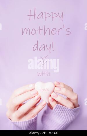 Grußkarte zum Muttertag. Weibliche Hände halten Keramik rosa Herz mit Liebe auf dem weichen violetten Hintergrund Stockfoto