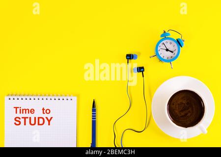 Blauer Wecker, blaue Kopfhörer, Tasse Kaffee und Notizbuch mit blauem Stift, liegt auf hellem gelben Hintergrund.Studien- und Lernkonzept. Kopierbereich. Stockfoto