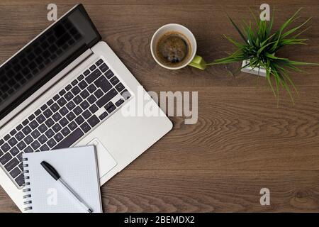 Ansicht von oben des Arbeitsbereichs. Tisch Schreibtisch mit Laptop, Kaffee, Notizblock, Stift und Pflanze mit Leerraum Hintergrund für Design. Flache Ansicht von dunkelbraunem Holz. Stockfoto