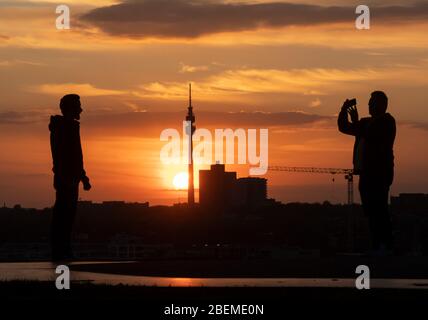 Dortmund, Deutschland. April 2020. Zwei junge Männer fotografieren sich bei Sonnenuntergang auf der Schlammhappe am Lake Phoenix. Im Hintergrund der Fernsehturm 'Florian', eines der Wahrzeichen der Stadt. Um das Corona-Virus einzudämmen, hat NRW alle Ansammlungen von drei oder mehr Menschen in der Öffentlichkeit verboten. Quelle: Bernd Thissen/dpa/Alamy Live News Stockfoto