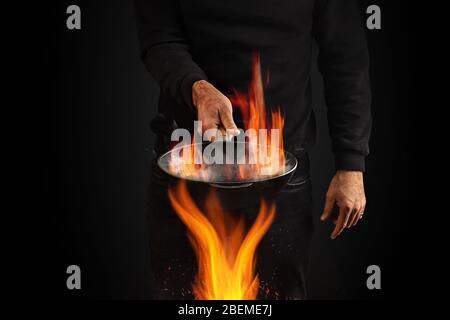 Junger Mann in Pullover und Jeans gekleidet. Er hält brennende Wok Pfanne mit Rauch über Feuer, vor schwarzem Studio Hintergrund. Kochkonzept. Nahaufnahme Stockfoto