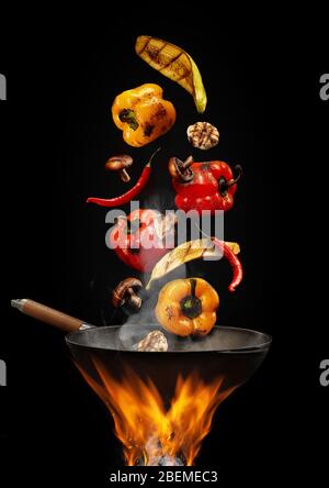 Wok Pfanne mit Rauch über Feuer ist Braten gelb und rot Paprika, Paprika, Pilze, Zucchini, Knoblauch vor schwarzem Studio Hintergrund. Nahaufnahme Stockfoto