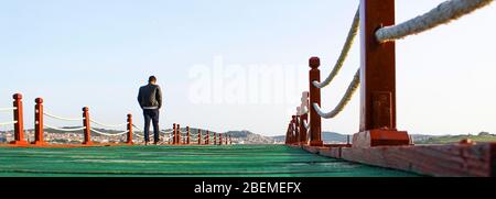 Mann, der alleine auf dem Pier läuft. Das Skelett ist aus Holz. Zaun aus Holzpfosten und Seil. Das Foto wurde bei bewölktem Wetter aufgenommen. Stockfoto