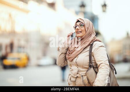 Muslimische Frau mittleren Alters trägt Hijab mit einem glücklichen Gesicht in der städtischen Umgebung stehend, auf ihrem Smartphone. Stockfoto