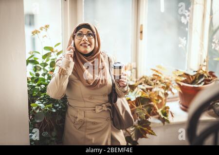 Muslimische Frau mittleren Alters, die Hijab mit einem glücklichen Gesicht trägt, steht am Fenster und nimmt ihr Smartphone auf. Stockfoto