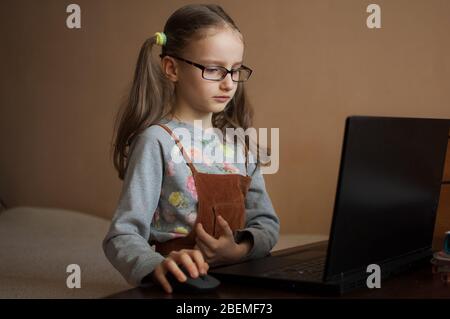 Nettes kleines Mädchen in eyeglassess sitzt vor ihrem schwarzen Laptop und studiert zu Hause wegen der Pandemie von Coronavirus Covid-19 und selbst Stockfoto