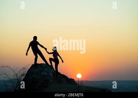 Mann und Frau Wanderer helfen einander, einen großen Stein bei Sonnenuntergang in den Bergen zu klettern. Pärchen klettern auf einem hohen Felsen in der abendlichen Natur. Tourismus, Reisen Stockfoto