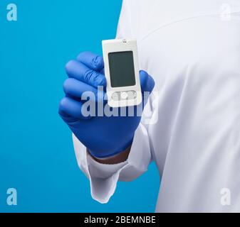 Die männliche Hand mit blauen sterilen Handschuhen hält ein weißes Blutzuckermessgerät zur Messung von Blutzuckerwert und blauem Hintergrund Stockfoto