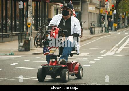 New York, NY, USA. April 2020. Ein Mann trägt eine Maske und einen NY Yankees Hut, während er am 14. April 2020 in New York auf einem mobilisierten Rollstuhl die Neunte Avenue in Chelsea auffährt. Kredit: Bryan Smith/ZUMA Wire/Alamy Live News Stockfoto