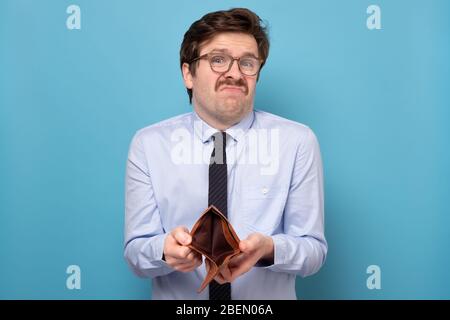 Trauriger Geschäftsmann, der eine leere Brieftasche auf blauem Hintergrund isoliert hält. Konkurs finanzielle Schwierigkeiten Konzept. Jobverlust. Stockfoto