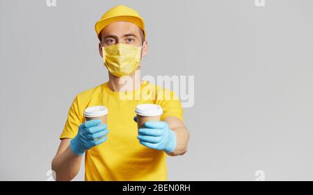 Paket an die Tür. Kurier in gelber Uniform, Schutzmaske und Handschuhe liefert Kaffee zum Mitnehmen. Lieferservice unter Quarantäne. Platz für Text Stockfoto