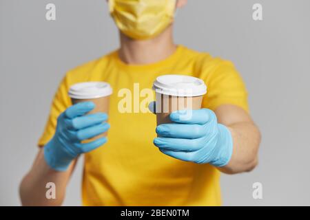 Paket an die Tür. Kurier in gelber Uniform, Schutzmaske und Handschuhe liefert Kaffee zum Mitnehmen. Lieferservice unter Quarantäne. Stockfoto