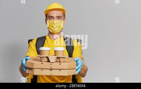 Paket an die Tür. Kurier in gelber Uniform, Schutzmaske und Handschuhe liefert Pizza und Kaffee zum Mitnehmen. Lieferservice unter Quarantäne. Kopieren Stockfoto