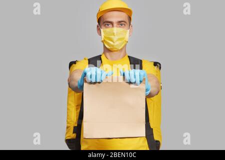 Kurier in gelber Uniform, Schutzmaske und medizinische Handschuhe liefern Essen und Kaffee zum Mitnehmen. Lieferservice unter Quarantäne. Kopieren Sie den Bereich für den tex Stockfoto