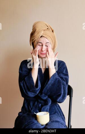 Frau in einem Gewand und mit einem Handtuch auf dem Kopf trägt Kosmetik auf die Haut Stockfoto