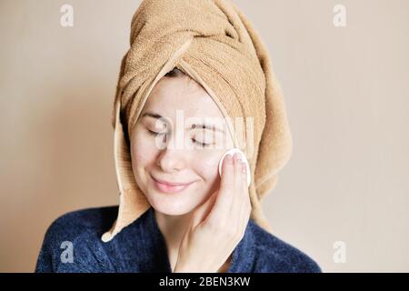 Frau in einem Gewand und mit einem Handtuch auf dem Kopf trägt Kosmetik auf die Haut Stockfoto