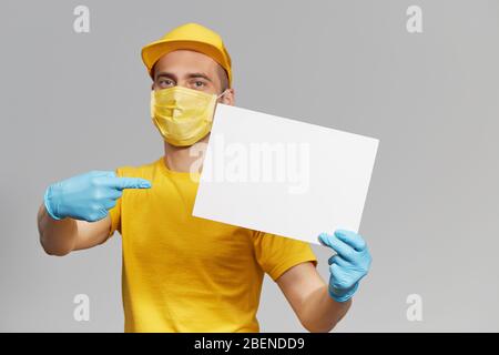 Kurier in gelber Uniform, Schutzmaske und Handschuhe liefert Essen und Kaffee zum Mitnehmen. Lieferservice unter Quarantäne. Halten Sie ein weißes Schild für t Stockfoto