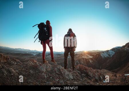 Ein paar junge Wanderer mit großen Rucksäcken stehen und schauen auf Berge und Sonnenuntergang Stockfoto
