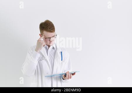 Young Trainee Arzt Praktikant steht auf weißem Hintergrund. Schöner rothaariger Mann, denkend Stockfoto