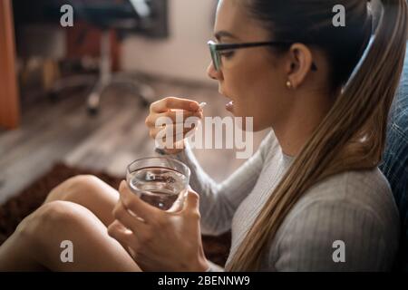 Seitenansicht EINER jungen kranken weißen Frau zu Hause schlucken ihre medizinische Behandlung Pillen mit einem Glas Wasser. Stockfoto