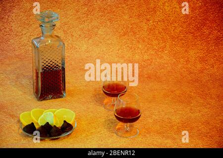 Zwei Gläser und eine Kristallflasche mit Cognac stehen neben einem Teller mit Zitronen- und Schokoladenzuckern auf braunem Hintergrund. Nahaufnahme. Stockfoto