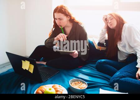 Eine Schülerin arbeitet mit einem Laptop, die zweite junge Frau schläft ein. Zwei rothaarige Studenten studieren zu Hause oder in einem Studenten Stockfoto