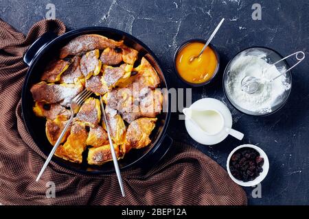 Österreichischer Süßpfannkuchen kaiserschmarrn mit Apfelmus, Rosinen und Puderzucker, goldenes Besteck auf einer schwarzen Backform auf dunklem Betongrund Stockfoto