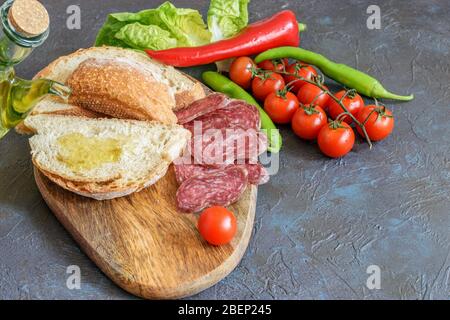 In Scheiben geschnittenes Fuet, Olivenöl tropft auf das Weißbrot. Etwas Pfeffer, Grün und Tomaten Kirsche auf dem dunklen Hintergrund. Gesunde mediterrane Küche. Stockfoto