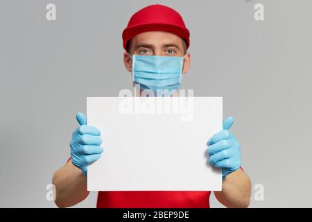 Kurier in roter Uniform, Schutzmaske und Handschuhe mit einem weißen Schild für Text. Lieferservice unter Quarantäne. Stockfoto