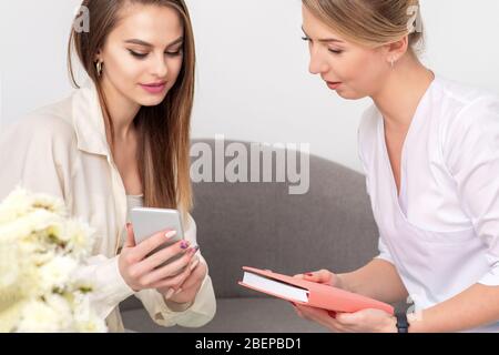 Kosmetologe spricht mit jungen Frau im Bademantel Blick auf das Smartphone des Kunden. Stockfoto