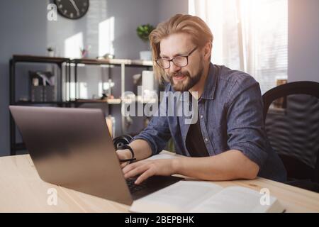 Fröhlicher bärtiger Mann in Denim-Kleidung, der am Laptop arbeitet, während er am Tisch sitzt. Reife männliche mit trendy Haarschnitt Gefühl glücklich während Remote-Arbeit bei h Stockfoto
