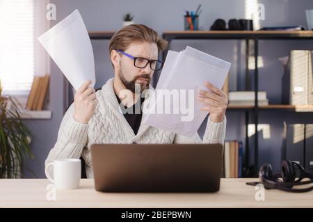 Konzentrierter Mann in lässigem Outfit und Brille, die Dokumente liest, während er mit geöffnetem Laptop am Tisch sitzt. Bärtige Person, die während der STA Stockfoto