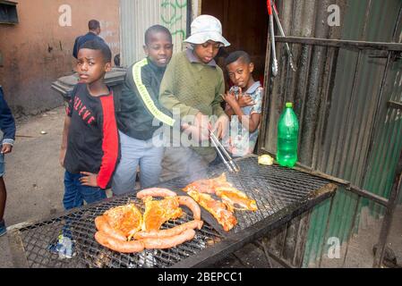 Jungs kochen Fleisch auf einem Grill in der Seitengasse eines Townships in Kapstadt. Sie sind zuversichtlich und es gibt eine gute Auswahl an Fleisch. Hütten umgeben die Gasse in der Langa Township. Die Jungs sind gut gefüttert, gut gekleidet und für das Kochen zuständig. Sie nennen ein BBQ ein Braai in Südafrika und diese Art der Küche ist sehr beliebt. Stockfoto