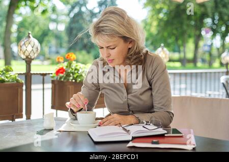 Charmante blonde Frau mittleren Alters mit einer Tasse Kaffee im Café im Freien, reife Geschäftsfrau in der Kaffeepause Stockfoto