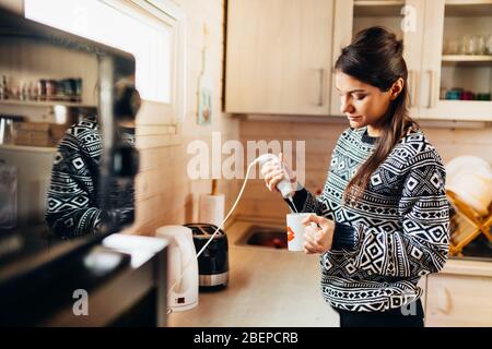 Frau, die Kaffee trinken zu Hause Küche mit elektrischen Milchaufschäumer Getränk Mixer Kaffee Schneebesen für schaumige Sahne Schlagkaffee.Cafe Latte.Self-isolati Stockfoto