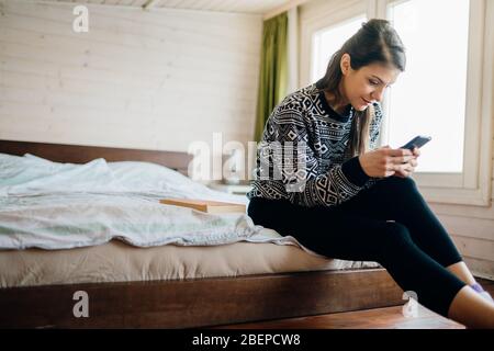 Junge Frau, die Nachrichten auf dem Handy vom Bett aus eintippt.isolation.Communicating in Quarantäne bleiben.Lachende Frau, die soziales Netzwerk nutzt Stockfoto