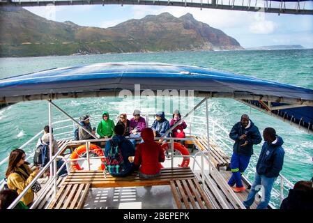 Touristen sitzen auf dem Deck eines Bootes und fahren zur Seal Island in Hout Bay, nahe Kapstadt, Südafrika. Das Meer ist rau, bedingt durch windige Bedingungen. Zwei Wachen stehen auf dem Deck, um die Besucher zu schützen. Die Touristen kommen aus Japan und wickeln sich gut gegen den starken Wind ein. Stockfoto