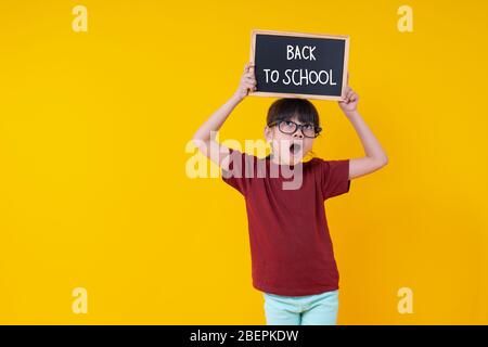 Junge asiatische Thai Mädchen Schüler, die kleine Tafel über Kopf mit zurück zu Schule Worte, wow und überrascht Kind Konzept auf gelben Bakkground in Gestüt Stockfoto