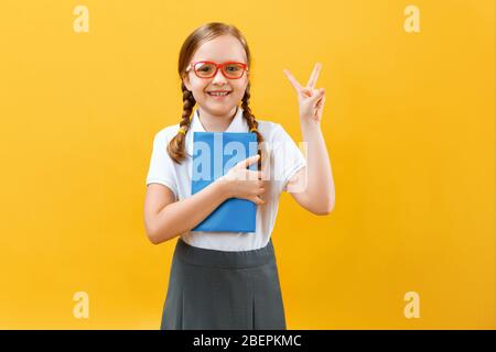 Porträt einer kleinen Schülerin mit Brille auf gelbem Hintergrund. Das Kind hält ein Buch in der Hand und zeigt ein Siegeszeichen. Bildung, Erfolg Stockfoto