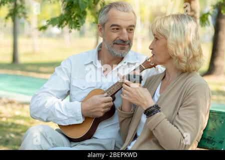 Glückliches älteres Paar sitzen spielen Ukulele und trinken Kaffee im Park Stockfoto