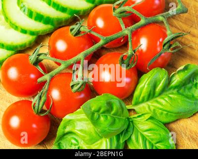 Vittoria Kirschtomaten auf der Rebe mit Gurkenscheiben und frischen Basilikumblättern auf einem Holzhackbrett Stockfoto