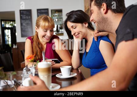 Drei Freunde schauen sich Fotos auf dem Handy im Café an Stockfoto