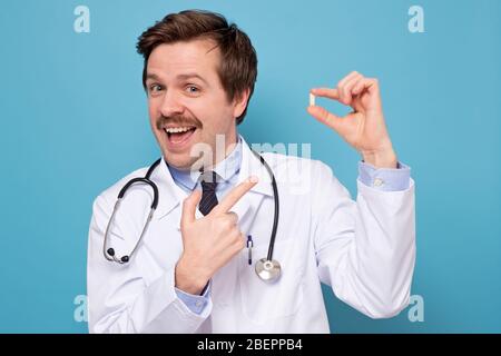 Fröhlich aufgeregt jungen Mann mit Schnurrbart stehen und zeigen auf Pillen. Studio auf blauer Wand gedreht. Stockfoto