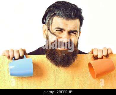 Bärtiger Mann, langer Bart. Brutaler kaukasischer weinender unrasiert Hipster mit orangefarbenem Papierblatt, das bunte Tassen im Studio-Hintergrund hält Stockfoto